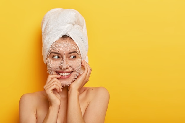 Retrato de mulher europeia aplica máscara facial orgânica para limpar a pele, cuida da tez, sorri gentilmente, mostra os dentes brancos, tem ombros nus, fica encostada na parede amarela com espaço em branco