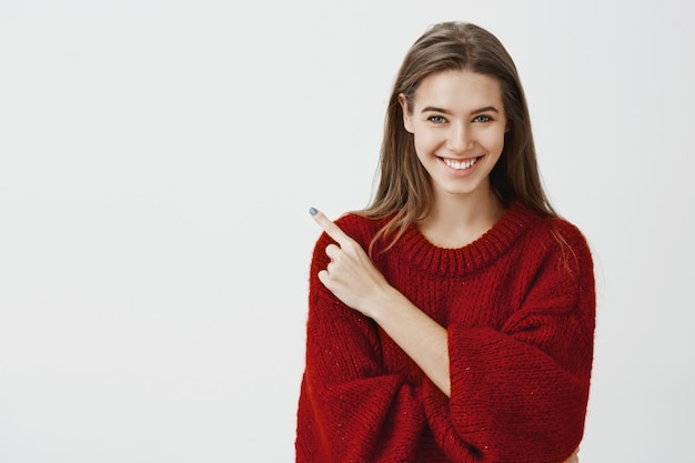 Retrato de mulher europeia encantadora positiva na elegante camisola solta vermelha, apontando no canto superior esquerdo e sorrindo amigável, dizendo bons conselhos ou item de publicidade perto de fundo cinza