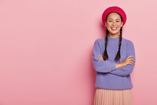 Retrato de mulher feliz com aparência oriental, com as mãos cruzadas sobre o peito, usa boina vermelha, blusa e saia roxas, posa contra a parede rosa, tem aparência entusiasmada