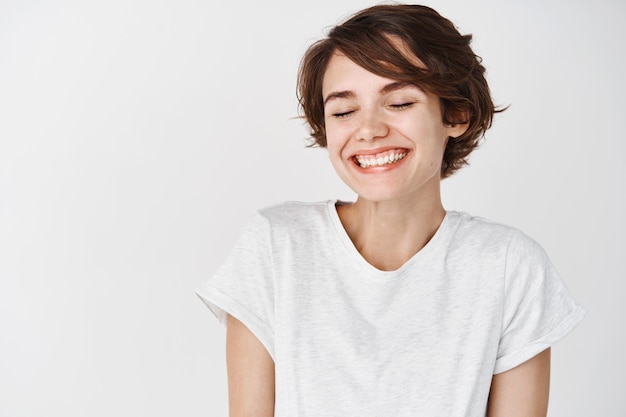 Retrato de mulher feliz e positiva fecha os olhos, sorrindo despreocupada, em pé com uma camiseta na parede branca