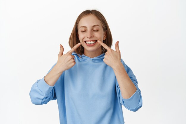 Retrato de mulher feliz mostrando os dentes brancos após o clareamento dentário, apontando o dedo para um sorriso perfeito, em pé com camiseta azul encostada na parede branca