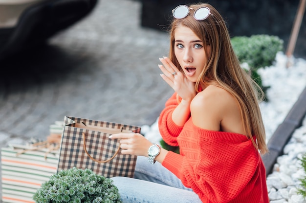 Foto grátis retrato de mulher feliz, mostrando roupas compradas em loja. ela está sentada na rua perto da boutique.