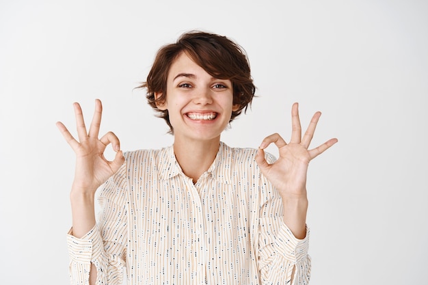 Retrato de mulher feliz natural com penteado curto, mostrando gestos bem e sorrindo, aprovar e gostar de algo, mostrar feedback positivo, parede branca