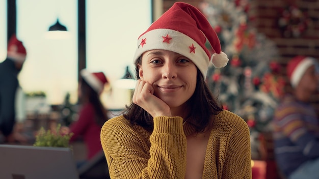 Retrato de mulher festiva sentada à mesa no escritório decorado, usando chapéu de Papai Noel para comemorar o Natal com colegas no local de trabalho. Trabalhando no pc durante a celebração do feriado sazonal.