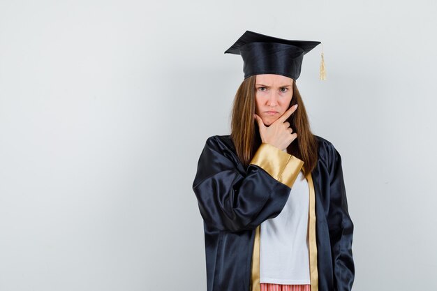 Retrato de mulher graduada segurando a mão no queixo em roupas casuais, uniforme e olhando malvado vista frontal