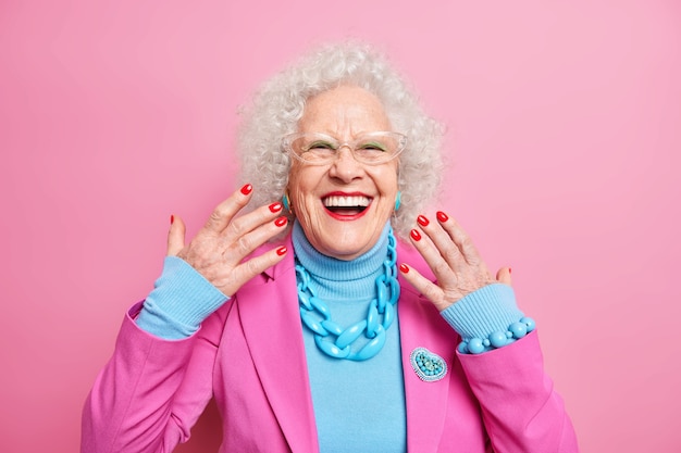 Retrato de mulher idosa de cabelo encaracolado levanta as mãos, sorri, usa óculos, colar de roupa da moda e está de bom humor