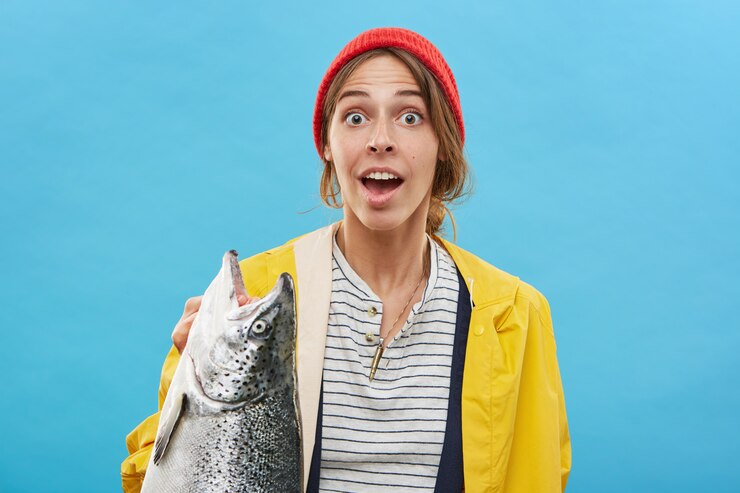 Foto grátis retrato de mulher jovem animado feliz em pé na parede azul em branco, segurando grandes peixes de água doce, sentindo-se alegre e espantado. conceito de pessoas, hobby, atividade, lazer e recreação