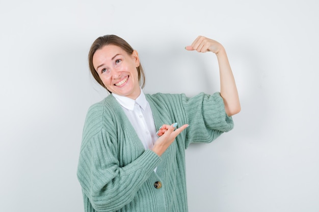 Retrato de mulher jovem apontando para os músculos do braço na blusa, casaco de lã e olhando a vista frontal orgulhosa