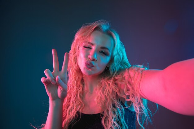 Retrato de mulher jovem caucasiana em fundo gradiente em luz de néon. Bela modelo feminino com aparência incomum. Conceito de emoções humanas, expressão facial, vendas, anúncio. Fazendo selfie, posando.