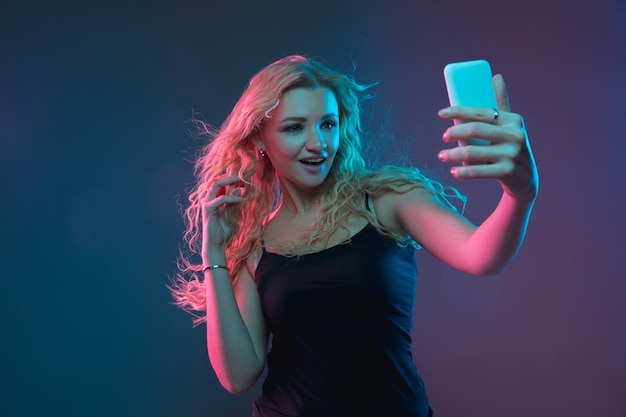 Retrato de mulher jovem caucasiana em fundo gradiente em luz de néon. Linda modelo feminino com aparência incomum. Conceito de emoções humanas, expressão facial, vendas, anúncio. Fazendo selfie, aposta, compra.