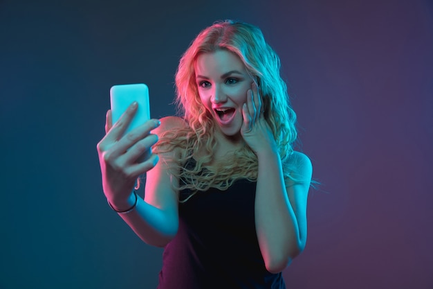 Foto grátis retrato de mulher jovem caucasiana em fundo gradiente em luz de néon. linda modelo feminino com aparência incomum. conceito de emoções humanas, expressão facial, vendas, anúncio. fazendo selfie, aposta, compra.