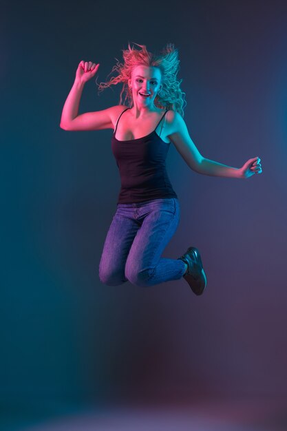 Retrato de mulher jovem caucasiana em fundo gradiente em luz de néon. Linda modelo feminino com aparência incomum. Conceito de emoções humanas, expressão facial, vendas, anúncio. Pulando, sorrindo.