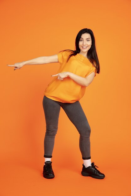 Retrato de mulher jovem caucasiana em fundo laranja do estúdio. Bela morena modelo feminino na camisa. Conceito de emoções humanas, expressão facial, vendas, anúncio. Copyspace. Apontando, mostrando.