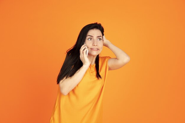 Retrato de mulher jovem caucasiana em fundo laranja do estúdio. Bela morena modelo feminino na camisa. Conceito de emoções humanas, expressão facial, vendas, anúncio. Copyspace. Falando ao telefone.