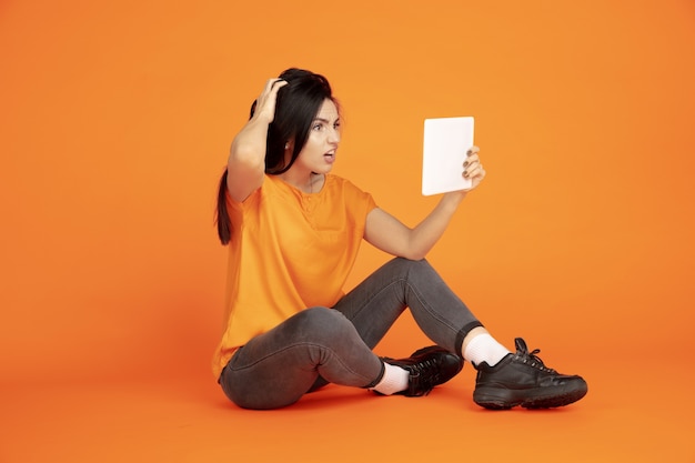 Retrato de mulher jovem caucasiana em fundo laranja do estúdio. bela morena modelo feminino na camisa. conceito de emoções humanas, expressão facial, vendas, anúncio. copyspace. usando tablet, vlogging.