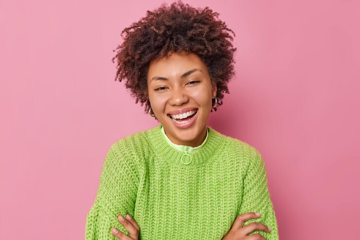 Foto grátis retrato de mulher jovem de cabelo encaracolado feliz sorri positivamente usa suéter de malha verde casual tem humor otimista expressa poses de emoções positivas contra fundo rosa ouve notícias agradáveis.
