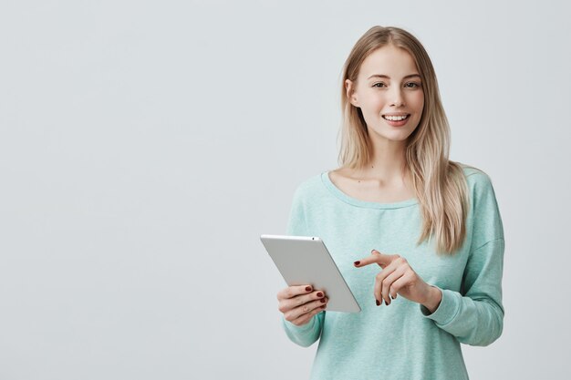 Retrato de mulher jovem e atraente com olhos escuros e cabelos loiros e compridos, vestindo blusa azul clara, trabalhando em tablet.