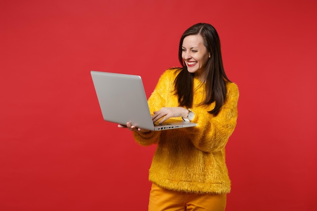 Retrato de mulher jovem e bonita sorridente no suéter de pele amarela, usando o computador laptop pc isolado em um fundo vermelho brilhante no estúdio. emoções sinceras de pessoas, conceito de estilo de vida. simule o espaço da cópia.