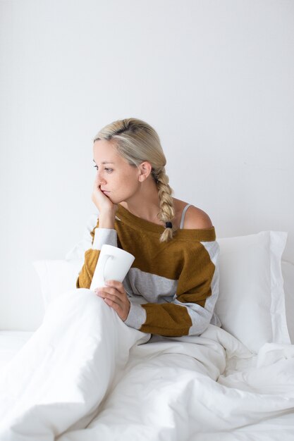 Retrato de mulher jovem e perturbada tomando chá na cama