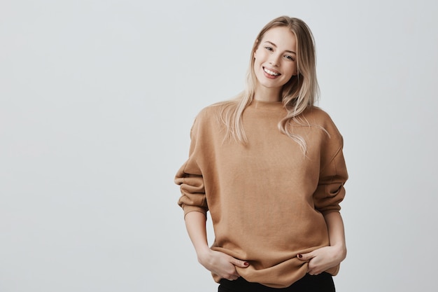 Foto grátis retrato de mulher jovem em roupas casuais com cabelos loiros tingidos, sorrindo suavemente durante uma conversa agradável, em pé na postura fechada