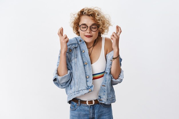 Retrato de mulher jovem esperançosa, atraente e elegante, desejando sorrir com os olhos fechados e expressões sonhadoras cruzar os dedos para dar boa sorte com as mãos levantadas, orando pela fortuna sobre a parede branca