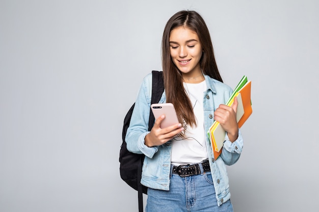 Retrato de mulher jovem feliz, de pé com a mochila, segurando livros e telefone celular isolado na parede branca