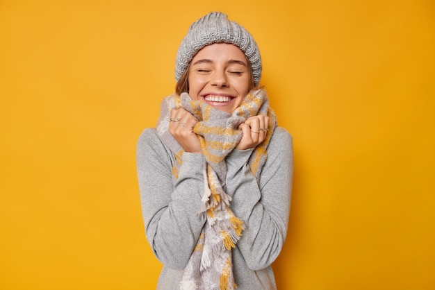 Retrato de mulher jovem feliz usa roupas quentes tricotada chapéu lenço em volta do pescoço fecha os olhos com satisfação sorri alegremente com dentes brancos isolados sobre fundo amarelo tem memória agradável