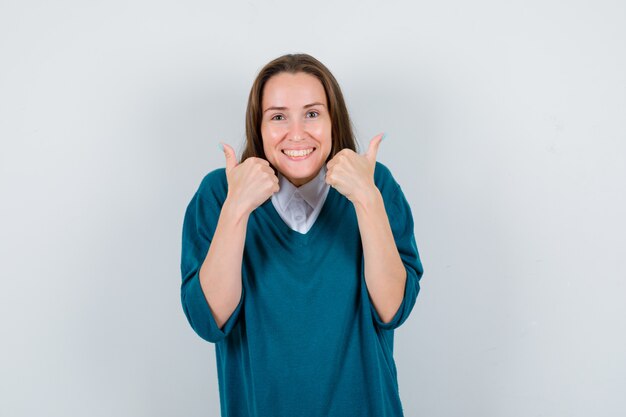 Retrato de mulher jovem mostrando dois polegares para cima com suéter sobre uma camisa branca e olhando frontalmente feliz