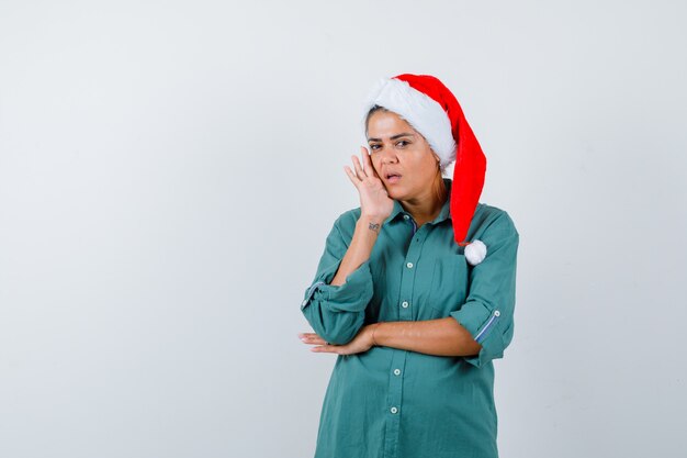 Retrato de mulher jovem segurando a bochecha com a camisa, chapéu de Papai Noel e olhando a vista frontal interessada