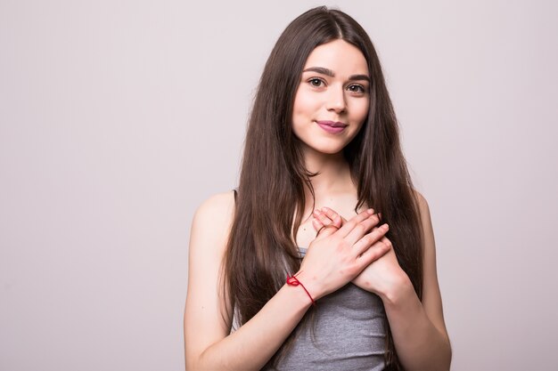 Retrato de mulher jovem sorridente positiva mantém as mãos no peito, expressa simpatia. Garota de bom coração, amigável, boa garota mostra bondade, vestindo uma camiseta cinza em branco na parede cinza