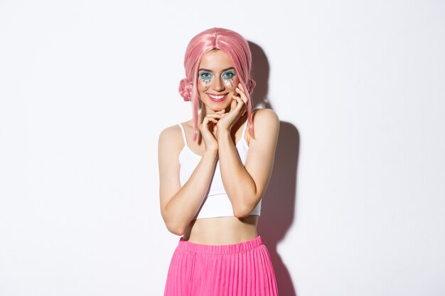 Retrato de mulher linda glamour com peruca rosa, maquiagem de halloween, sorrindo e parecendo coquete, em pé sobre um fundo branco.