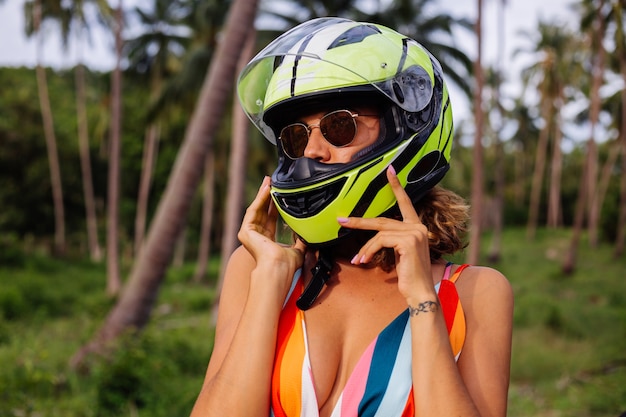 Retrato de mulher linda piloto no capacete verde amarelo da motocicleta e vestido de verão luz colorida na selva no campo tropical sob as palmeiras.