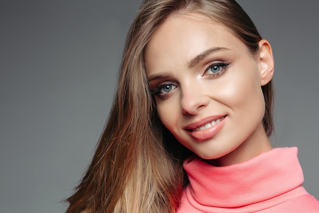 Retrato de mulher loira bonita e linda de suéter rosa com sensualidade de cabelo ondulado volumoso sorrindo para a câmera Garota com maquiagem perfeita após salão de beleza Fundo de estúdio cinza