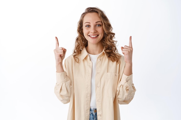 Retrato de mulher loira sorridente em roupas elegantes, apontando os dedos para cima no espaço da cópia, demonstra o texto promocional parecendo satisfeito com a câmera recomendando o fundo branco do produto