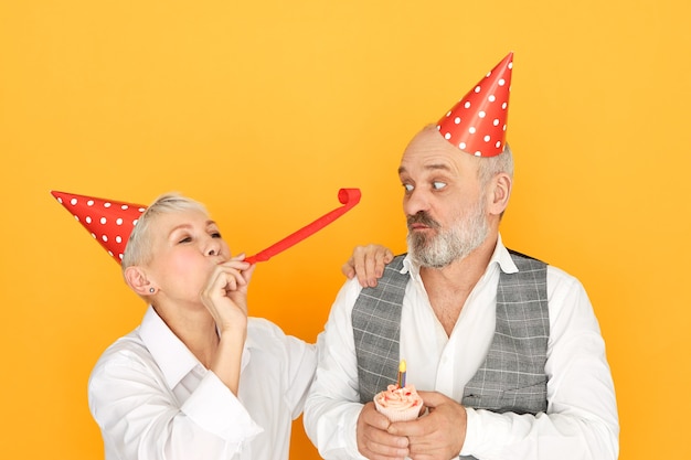 Foto grátis retrato de mulher madura alegre soprando tubo de papel enquanto se diverte na festa de aniversário ao lado de seu marido barbudo idoso com chapéu de cone