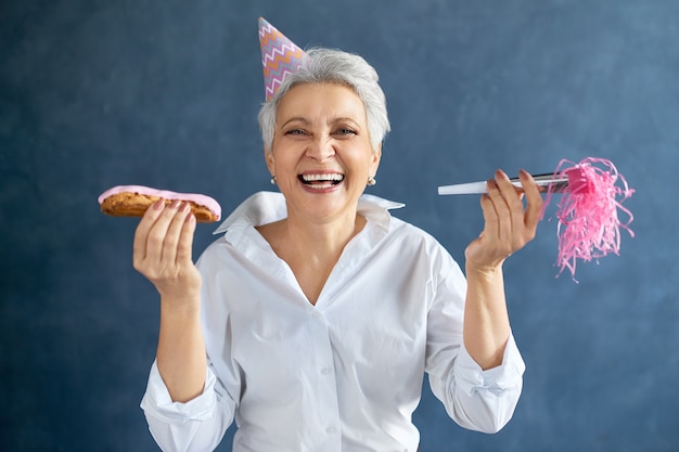 Retrato de mulher madura de cabelos grisalhos com chapéu de festa rosa segurando um apito e uma eclair