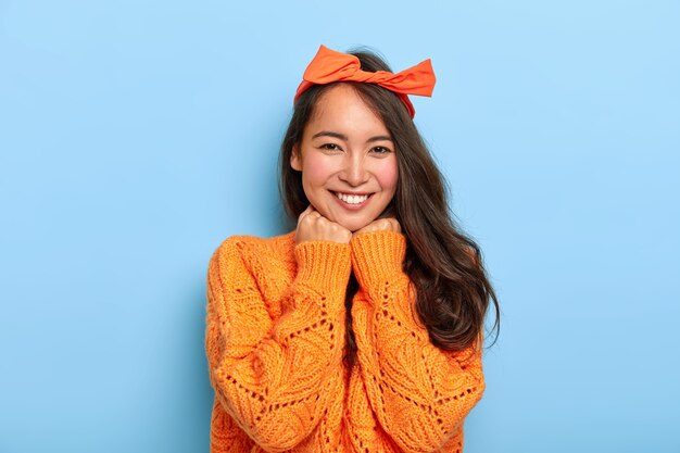 Retrato de mulher milenar morena feliz mantém ambas as mãos sob o queixo, sorri agradavelmente, usa tiara laranja e suéter de malha laranja quente