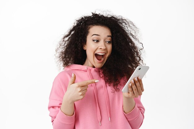 Retrato de mulher morena animada grita de alegria, apontando e olhando para a tela do celular, verificando a incrível oferta promocional on-line, de pé contra um fundo branco