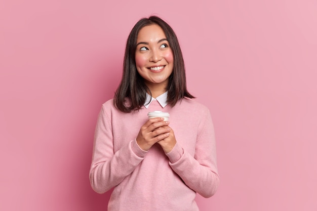Retrato de mulher morena asiática com expressão sonhadora sorri agradavelmente devaneios com café para viagem usa poses de jumper rosa puro interior. Modelo feminino pensativo se sente bem
