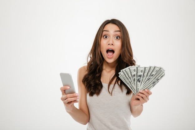 Retrato de mulher morena atraente 30 anos ganhando muito dinheiro dólar moeda usando seu smartphone, sendo alegre sobre branco
