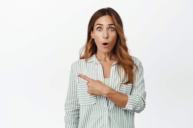 Foto grátis retrato de mulher morena impressionada na blusa, suspiro dizer wow, apontando o dedo para a esquerda no banner promocional de venda, chocado com grande desconto, fundo branco