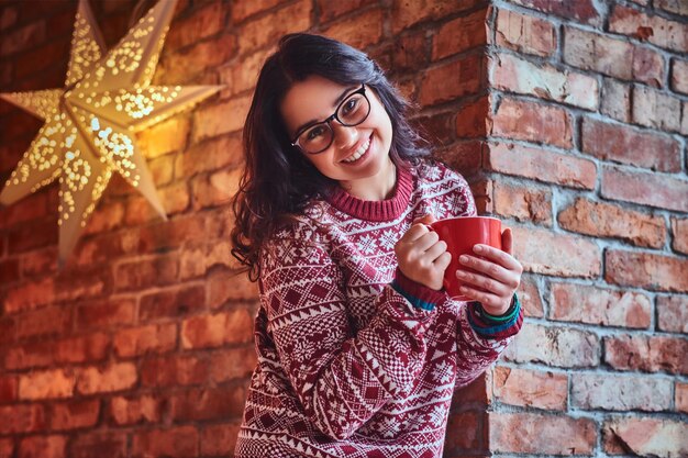 Retrato de mulher morena vestida com um suéter vermelho bebe café por cima da parede de um tijolo.