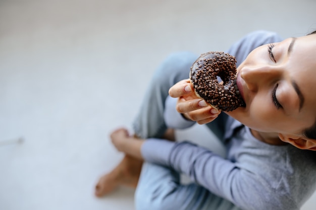 Retrato de mulher regozijando-se come rosquinha saborosa em casa.