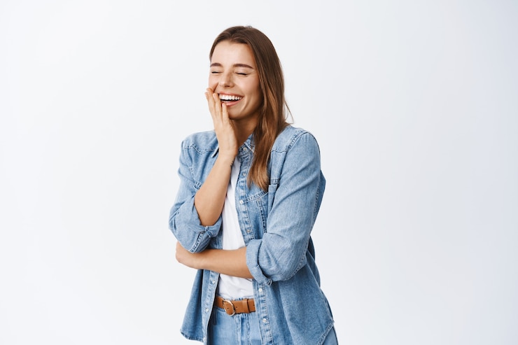 Foto grátis retrato de mulher rindo, sentindo-se feliz, se divertindo, fecha os olhos e ri de uma piada engraçada, em pé com roupas casuais contra uma parede branca