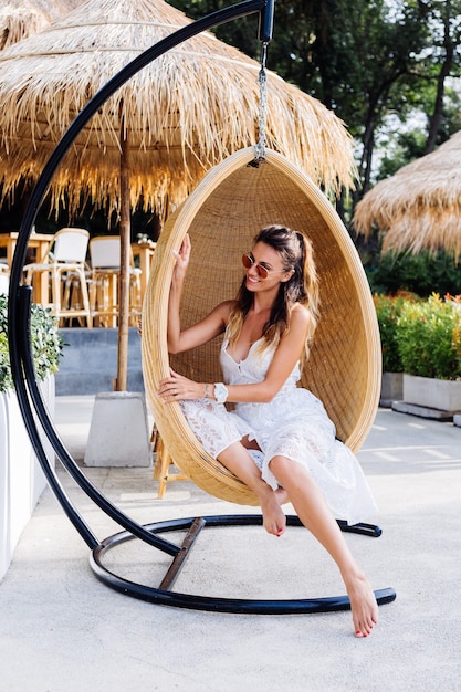 Retrato de mulher romântica europeia com vestido branco de verão fora do resort hotel em cadeira de palha de ovo.