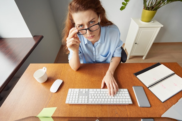 Retrato de mulher ruiva de camisa azul sentada em sua mesa em frente ao pc