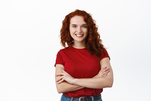 Retrato de mulher ruiva feliz e determinada com cabelo encaracolado, braços cruzados sobre o peito e sorrindo confiante, em pé com uma camiseta vermelha contra uma parede branca