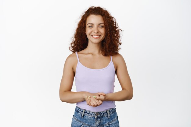 Retrato de mulher ruiva sorridente, olhando com expressão de rosto amigável e educada, de mãos dadas, ouvindo e tentando ajudar, auxiliando, de pé sobre fundo branco