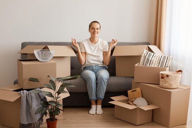 Retrato de mulher satisfeita sorridente, vestindo camiseta branca e jeans, sentado na tosse em um novo apartamento depois de se mudar, olhando para a câmera, posando em posição de mudra, cercado de pertences em caixas.