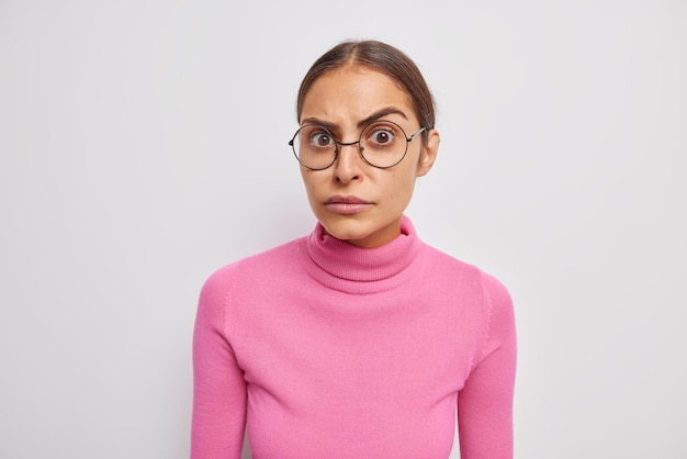 Retrato de mulher séria olha atentamente para a câmera e ouve algo surpreendente usa óculos redondos e poloneck rosa isolado sobre parede branca